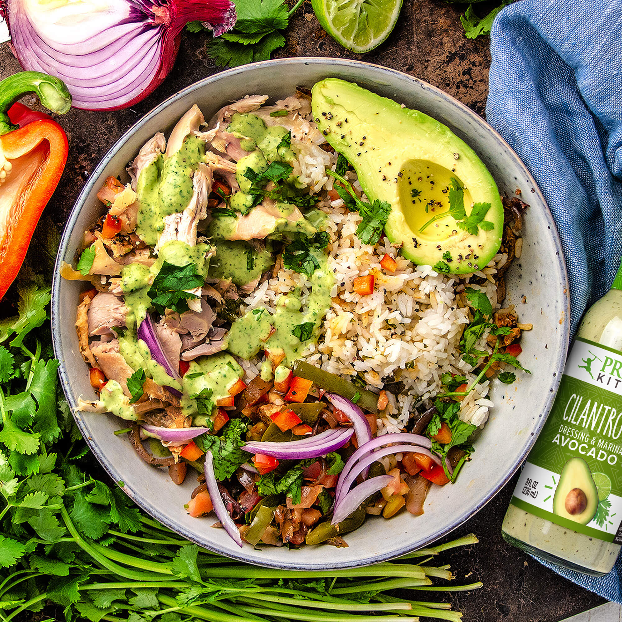 Mexican rice bowl with avocado and chicken, next to fresh herbs, veggies, and a bottle of Primal Kitchen Cilantro Lime creamy dressing. 
