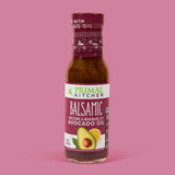 A bottle of Primal Kitchen Balsamic Vinaigrette and Marinade made with Avocado Oil with a dark purple label on a light pink background.