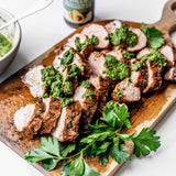 Juicy slices of pork with homemade chimichurri on a wooden cutting board. 