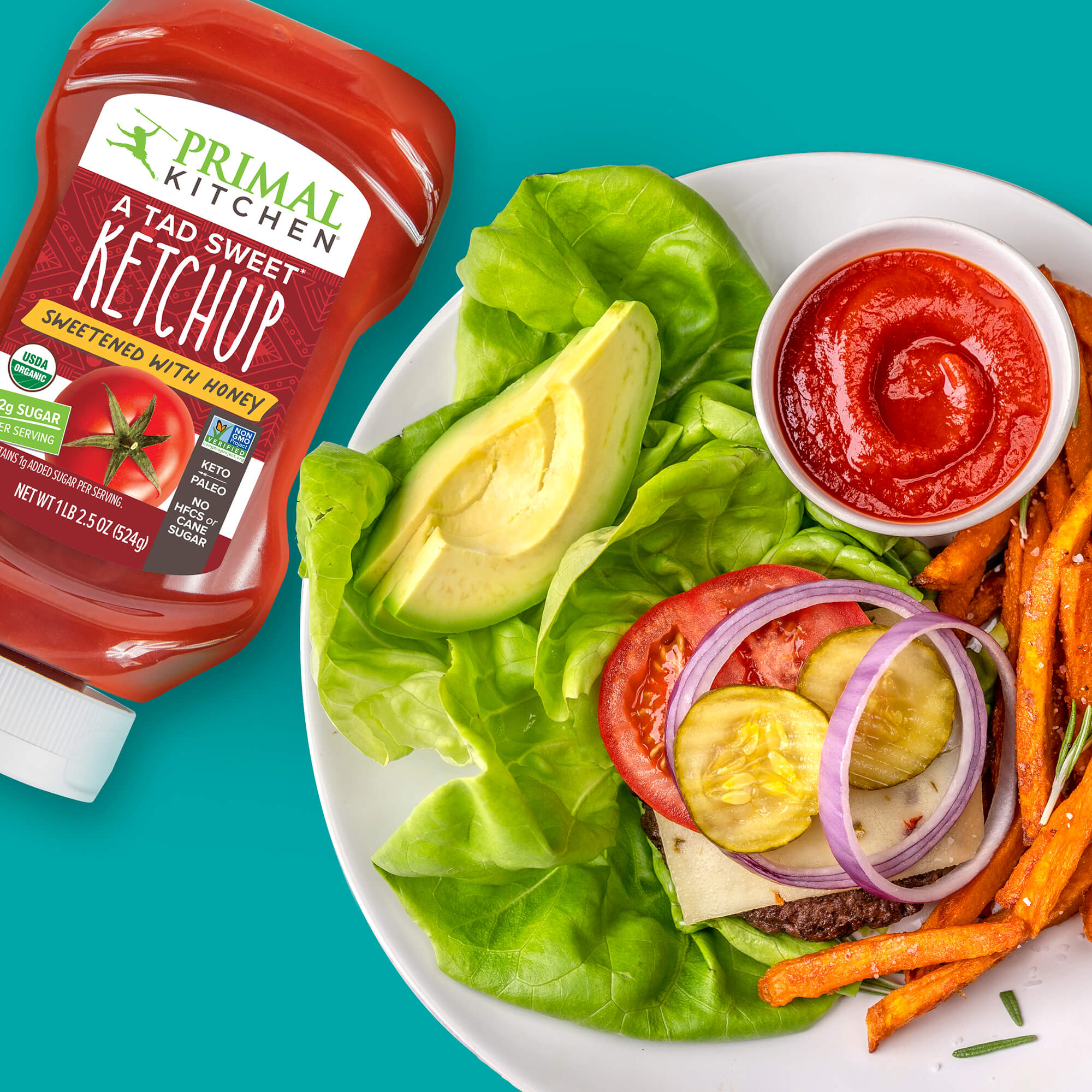 A Tad Sweet Squeeze Ketchup bottle beside an open face lettuce wrapped paleo burger with a side of sweet potato fries on a teal background.