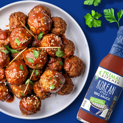 White plate with stacked beef meatballs topped with Korean BBQ Sauce, sesame seeds, and parsley with toothpicks, on a dark blue background with a bottle of Primal Kitchen Korean Style BBQ Sauce