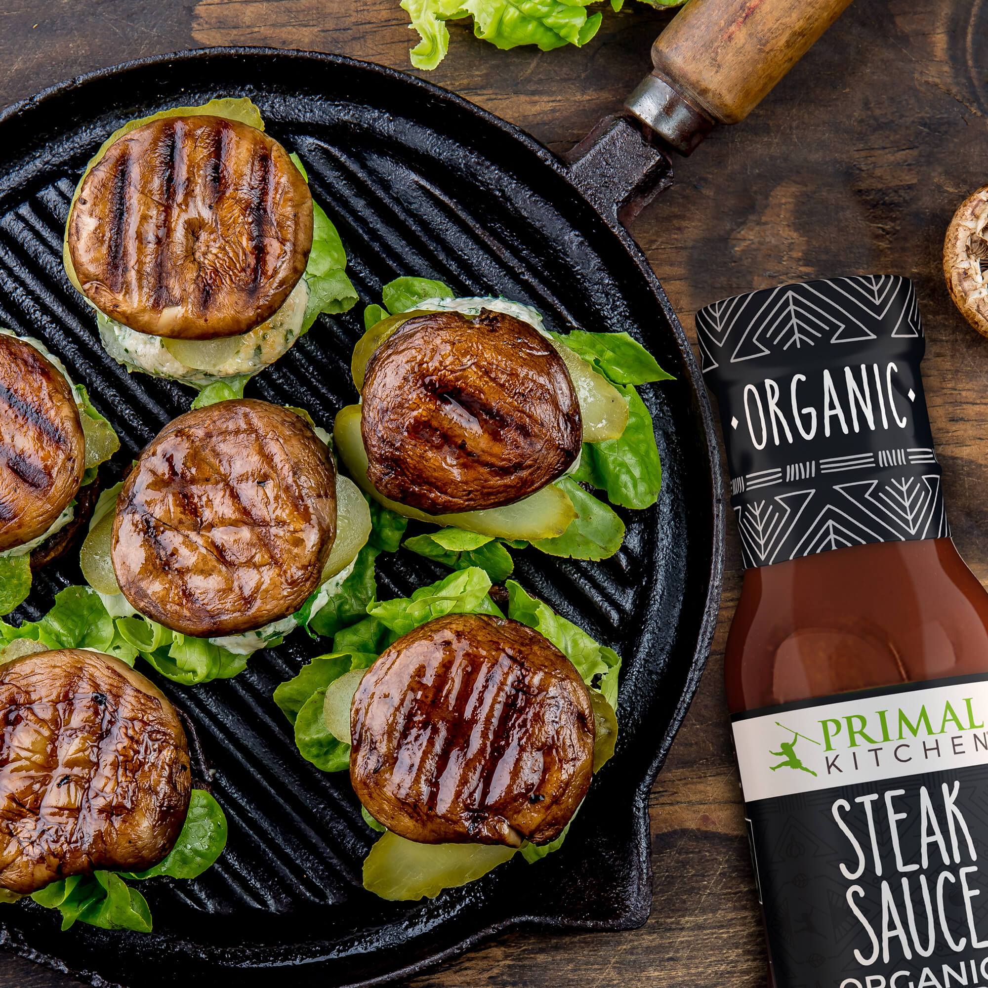 Grilled Portabella Mushroom burgers with organic lettuce and pickles on a skillet, alongside Primal Kitchen Organic Steak Sauce on a wood background