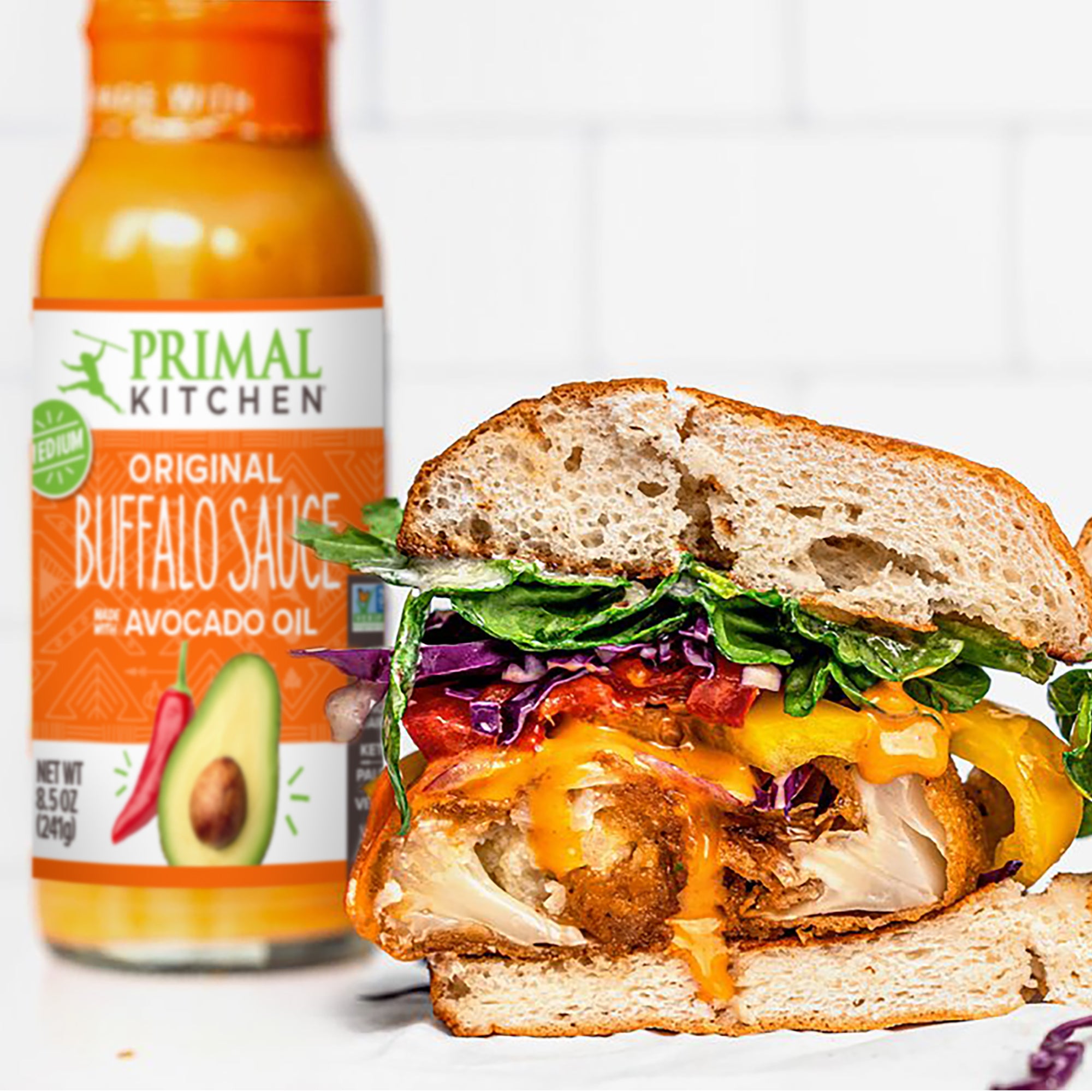 Bottle of Primal Kitchen Original Buffalo Sauce made with Avocado Oil next to a Buffalo Chicken Sandwich