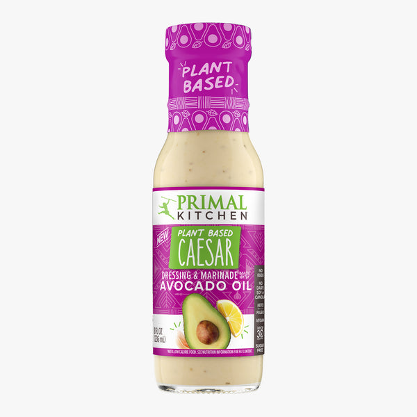 A bottle of vegan Plant Based Primal Kitchen Caesar Dressing & Marinade made with avocado oil and no eggs or dairy, with a hot pink-purple label, on a light grey background.