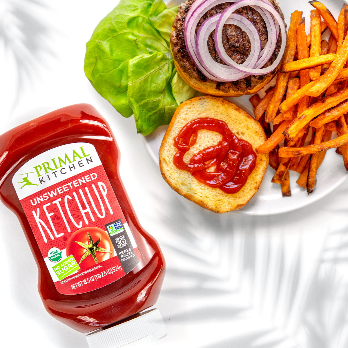 Primal Kitchen Unsweetened Ketchup in a squeeze bottle next to a plate with an open-faced paleo burger with lettuce, onions, and sweet potato fries