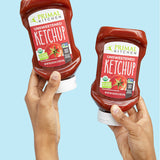 Two hands holding up Primal Kitchen Unsweetened Squeeze Ketchup bottles with No Added Sugar on a light blue background