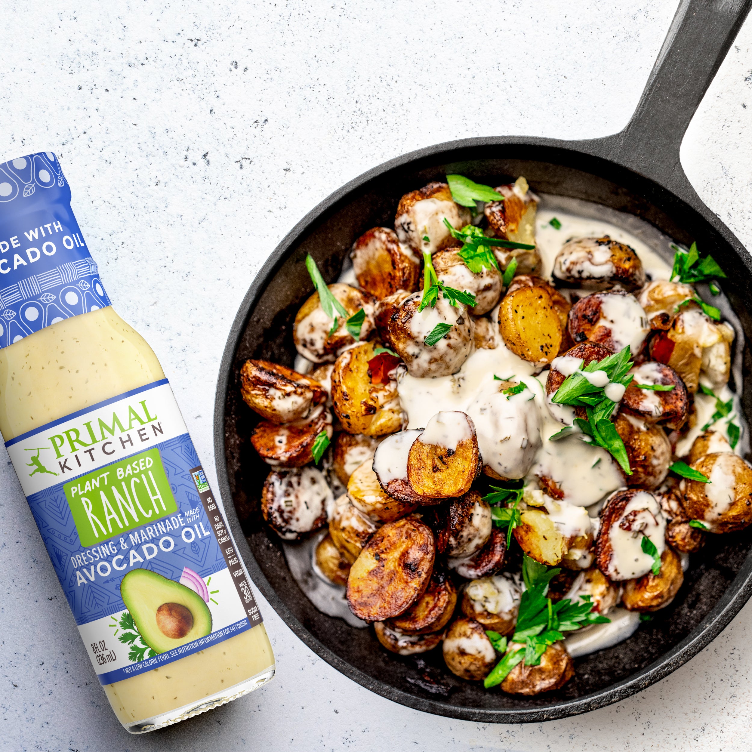 A skillet of golden brown sliced potatoes topped with a creamy vegan ranch sauce next to a bottle of Primal Kitchen Plant Based Ranch Dressing.